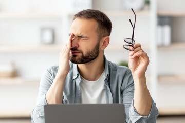 Poster - Caucasian male freelancer feeling tired, rubbing irritated eyes, sitting at desk with laptop, exhausted from online work. Young man having visual fatigue, doing business project on web