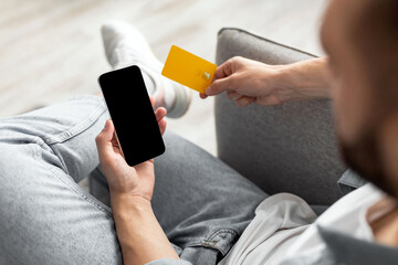 Poster - Unrecognizable millennial man with credit card and mobile phone shopping online from home, mockup for app or website. Closeup of young guy buying products in internet