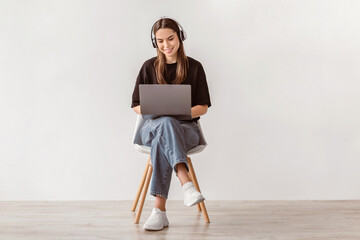 Poster - Cheery Caucasian woman in headphones having online video call on laptop, sitting on chair against white wall, full length. Millennial lady participating in webinar, communicating remotely on internet