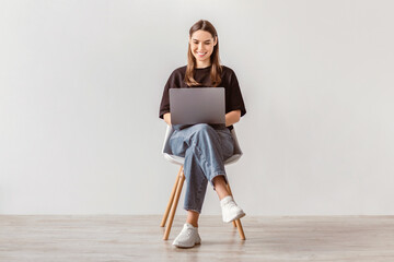 Wall Mural - Happy young woman working online, sitting on chair and using laptop against white studio wall, full length. Positive Caucasian lady surfing internet, watching webinar on portable computer