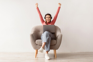 Poster - Excited young woman in headphones sitting in armchair, celebrating online win, great deal or business success. Millennial lady enjoying big sale in web store, feeling victorious