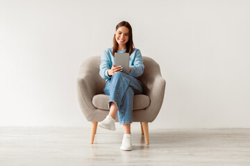 Poster - People, technology and communication concept. Portrait of smiling young woman with digital tablet sitting in armchair, working online against white studio wall, full length