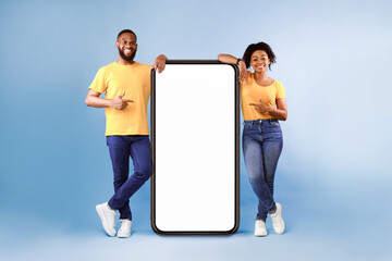 Poster - Great mobile app. African American couple pointing at giant smartphone with mockup, promoting application or website, advertising product or service, blue background. Full length