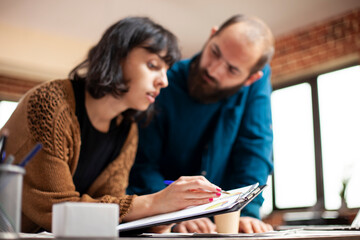 Wall Mural - Caucasian female analyst holding clipboard and explaining market analysis result to male coworker with beard. Closeup on businesswoman and man researching startup company financial documents at desk.