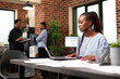 © DC Studio - Female manager sitting at table and using laptop in startup office. African american businesswoman surrounded with paperwork on desk, researching and comparing marketing trends on digital device.