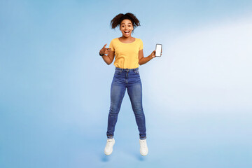Poster - Cool mobile app. Excited African American woman pointing at smartphone with blank screen and jumping over blue studio background. Free space for mockup, advertisement, website or application design