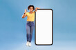 © Prostock-studio - New awesome mobile app. Positive African American lady posing near huge cellphone with empty screen, showing okay gesture, recommending website, presenting space for advertisement on blue