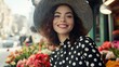 © Matt - Smiling woman with curly hair and a large black hat, surrounded by flowers in a vibrant outdoor market, wearing a polka dot dress
