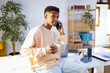 © Jimena Roquero/Stocksy - young man drinking tea while speaking on smartphone in bright office