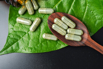 Wall Mural - Pile of herbal capsules on wooden spoon with herbs and green natural Betel pepper leaf background