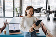 © Maria Vitkovska - Asian businesswoman smiling and holding clipboard in modern office with laptop showing graphs