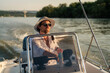 © DusanJelicic - Young man enjoying a leisurely sunset cruise on a river, skillfully navigating a motorboat while dressed in a stylish white shirt, sunglasses, and a fashionable hat