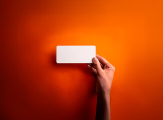 Aufkleber - A hand holds a blank white rectangular card against a vibrant gradient of orange. Dramatic lighting highlights the card in a studio setting, creating a focused presentation.