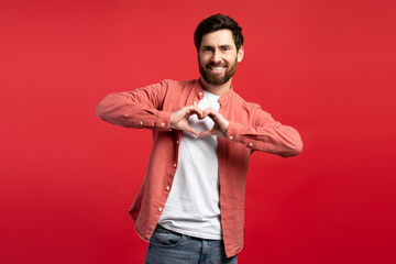 Wall Mural - Smiling young man showing heart gesture near chest, looking at camera, isolated on red