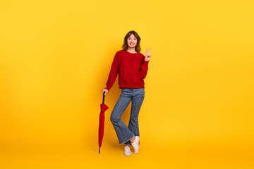 Young woman in casual attire holding a red umbrella while posing against yellow studio background making a peace gesture