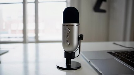 Poster - Silver microphone resting on white table, laptop with audio editing software blurred in background, capturing podcast production environment in smooth slow motion