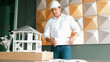 © Summit Art Creations - Portrait of professional project manager holding project plan with house model at table. Professional architect engineer wearing safety helmet while standing posing with confident. Manipulator.