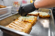 © pavel siamionov - A close-up of a gloved hand adding cheese and a sauce to slices of toasted bread in a commercial kitchen, showcasing food preparation for a delicious meal.