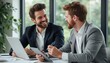 © natakot - The image captures two young adult men, one with a beard and the other clean-shaven, sharing a joyful moment while discussing ideas at a sleek office table. Their expressions radiate camaraderie and