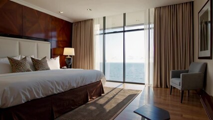 Poster - Sunlight illuminates a luxurious hotel room featuring a comfortable bed, an armchair, and a large window offering a breathtaking view of the ocean