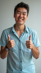 Canvas Print - A joyful young Asian man flashes a bright smile while giving two thumbs up, radiating positivity and confidence. His casual denim shirt and relaxed demeanor create an inviting atmosphere, perfect for