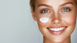 © Nati - A close-up portrait of a woman smiling as she gently applies face cream to her skin. Soft, natural lighting highlights her radiant expression against a bright, plain background.
