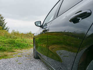 Wall Mural - Side of a black car and green field in front and reflection of a forest on a side of the car. Travel to nature concept. Grey cloudy sky.