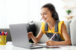 © Prostock-studio - Online Video Call And Communication. Portrait of cheerful teenage pupil in headset sitting on couch at table, using pc having virtual conference, talking with teacher or friends, writing holding pen