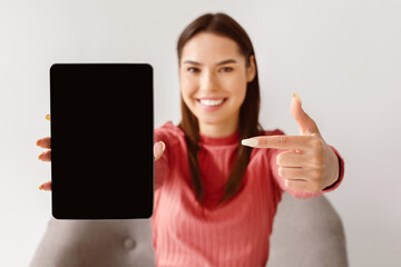 Poster - Happy Caucasian woman pointing at tablet pc with black blank screen for mockup, presenting new application, offering space for website, advertising your product or service, selective focus