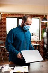 Wall Mural - Businessman wearing face mask and placing his laptop on table in brick wall room, ready for new day at work. Male employee standing at office desk and holding his personal computer in startup company.