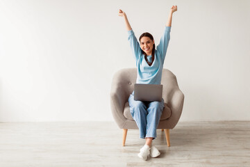 Poster - Triumphant Caucasian woman in armchair raising hands up, celebrating online win, enjoying sale in web store, using laptop computer against white studio wall, blank space