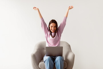 Wall Mural - Excited Caucasian lady with laptop raising her arms up, feeling overjoyed, winning online casino bet, shopping on huge discount, achieving success, sitting in armchair on white studio background