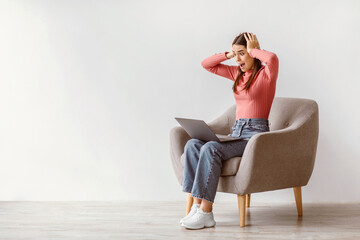 Poster - Deadline concept. Portrait of shocked Caucasian woman sitting in armchair with laptop computer and grabbing her head against white studio wall, copy space. Stressed young lady working online