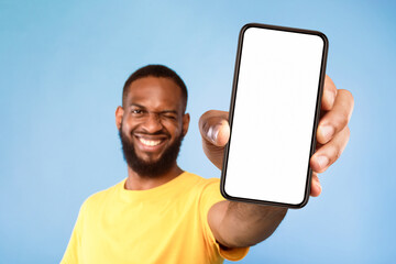 Wall Mural - Mobile app advertisement. Smiling young African American man showing smartphone with empty screen, winking on blue studio background, selective focus. Cellphone display mockup, space for ad or website