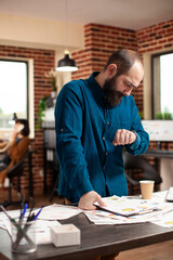 Wall Mural - Bearded man checking time and preparing for business project presentation in startup office. Portrait of male analyst looking at his wristwatch and standing at desk filled company research documents.