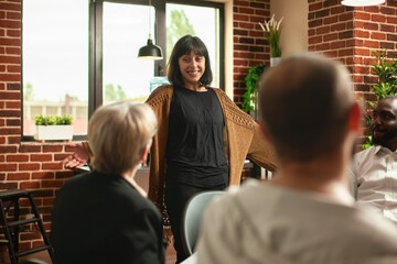 Smiling white woman standing in circle of people, discussing her progress at therapy session in supportive space. Young female patient excitedly shares her recovery achievement at aa group meeting.