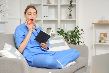 Wall Mural - Young female medical student reading book and eating apple on sofa at home