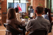 © DC Studio - Bearded man and coworkers discuss marketing plan, watching digital screen displaying female client on video call. Diverse team conduct business meeting with remote businesswoman visible on monitor.