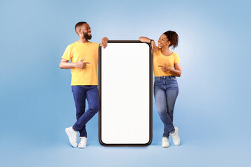 Poster - Hot offer, online sale. Funky African American young couple pointing at cellphone with mockup space over blue studio background. Guy and lady advertising new awesome mobile app or cool website