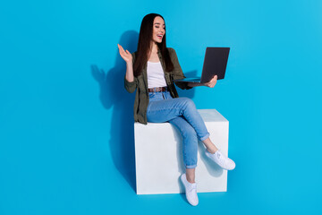 Wall Mural - Trendy young woman using a laptop while sitting on a white block with a blue background