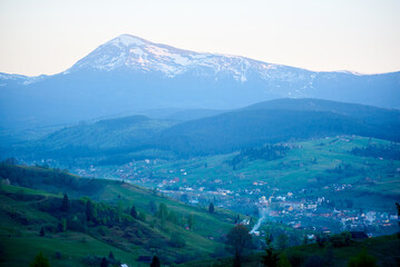 Scene of valley village surrounded by verdant hills, with majestic, snow-capped mountain in background. Early morning or late evening light casts serene glow, while wisps of smoke rise from chimneys.