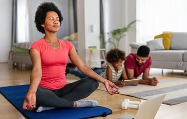 Wall Mural - young woman, mother exercising at home in living room, father playing with kid in background.