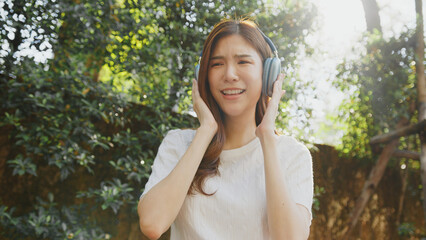 Wall Mural - Attractive Enjoying music outdoors, woman smiles while ing headphones in lush garden. sunlight creates warm atmosphere, enhancing her joyful expression. Asian Woman Lifestyle