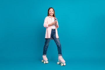 Wall Mural - Young girl roller skating with a phone against a vibrant blue background showcasing a happy and playful moment.