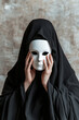 © Kassiopeia  - Dressed in all black, a figure stands against a textured backdrop, holding a white mask before their face. The dim lighting enhances the mysterious aura of the scene, creating a striking contrast