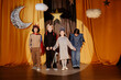 © Seventyfour - Young children standing on stage during a performance, holding hands and wearing costumes in front of a backdrop featuring stars and a crescent moon hanged from ceiling