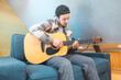 © Fauren - Young man with a relaxed demeanor sits barefoot on a cozy blue couch, strumming an acoustic guitar. His expression shows contentment and focus as he connects with his music. Casual and creative vibe