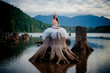© Connect Images - Woman in white gown with bow and arrow sits on massive tree stump by tranquil lake with forest and mountains. Rattlesnake Lake, Puget Sound, WA, USA