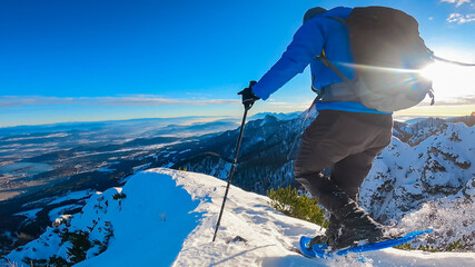 Wall Mural - Winter hiker with snowshoes and hiking poles stands on snowy summit of Mallestiger Mittagskogel in Carinthia, Austria, overlooking dramatic Karawanks mountain range during clear sunny winter day