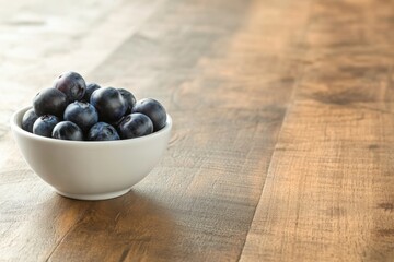 Aufkleber - A close-up view of fresh blueberries filled in a small white bowl placed on a wooden table, softly lit to enhance the healthy, inviting appearance